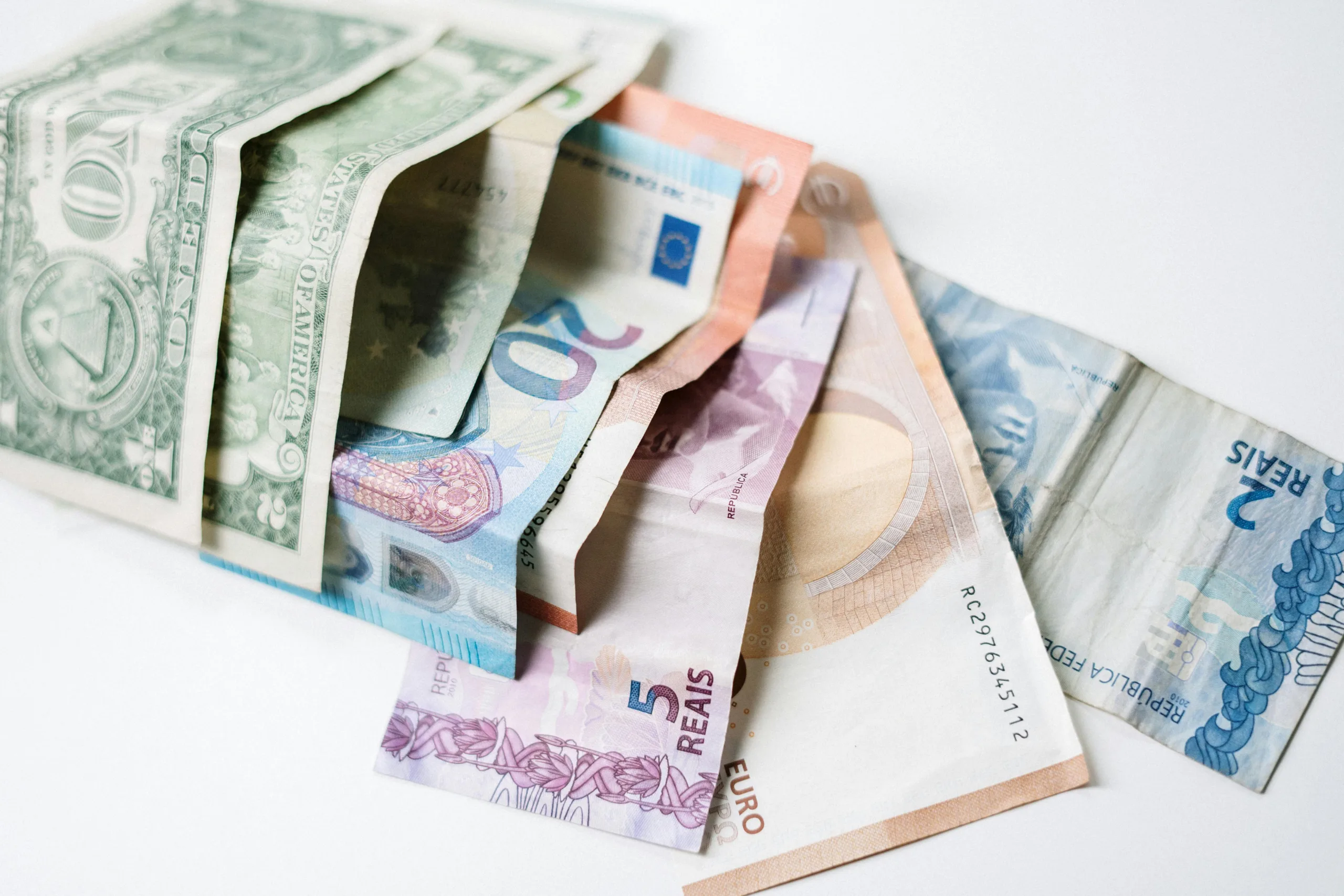 A collection of different international currency notes, including US Dollar, Euro, and Brazilian Real, on a white background.